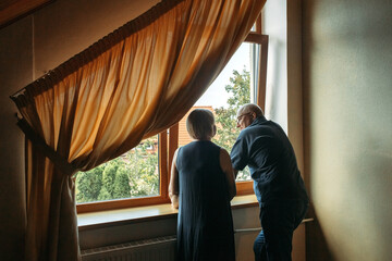 A happy elderly couple stand near the window, look.