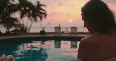 Girl silhouette near swimming pool at sunset time. Colorful sunrise sky reflected in calm water. Relax, retreat, meditation. Travel, tourism, holiday spa vacation.