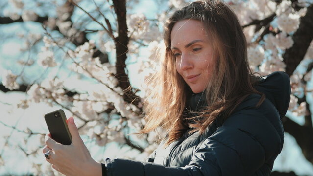 Girl making selfie with white sakura flowers in Tokyo, Japan. Woman enjoy spring blossom happy smiling looking to the camera. Sunny spring outdoors activity. Spring blossom of sakura tree in city park