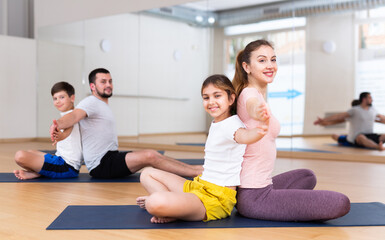 Fototapeta na wymiar Friendly sporty family of four practicing partner yoga in fitness studio. Teen girl doing exercises in pair with her mom..