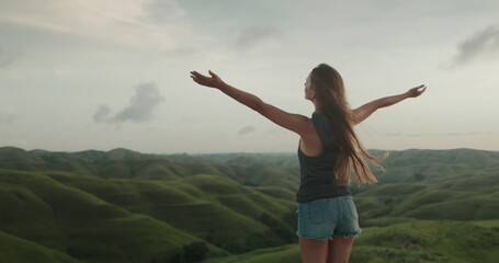 Woman enjoy sunset landscape, raises hands against green hills valley. Long hair brunette girl...