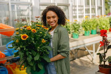 Cheerful woman gardener holding flower pot while standing on greenhouse yard background