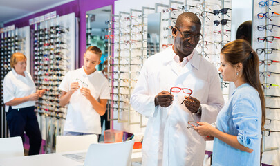 Professional smiling glad friendly African-American ophthalmologist consulting young woman looking for glasses at optics store - obrazy, fototapety, plakaty