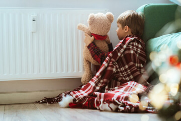 cute little boy wrapped id plaid sitting by heater warming teddy bear paws