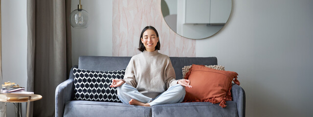 Relaxation and patience. Smiling young asian woman in cozy room, sitting on sofa and meditating,...