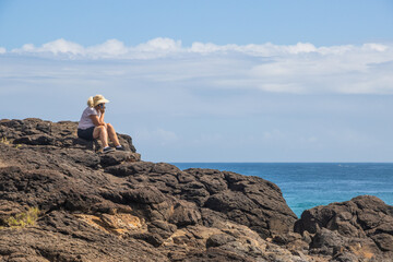 Woman sitting on a high cliff overlooking the ocean at Fingal Head on the East coast of Australia. She is on the phone.