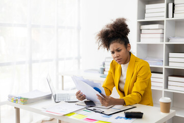 Beautiful African American black businesswoman working with laptop computer and financial documents at table in office typing documents, checking email, joining online meeting at work