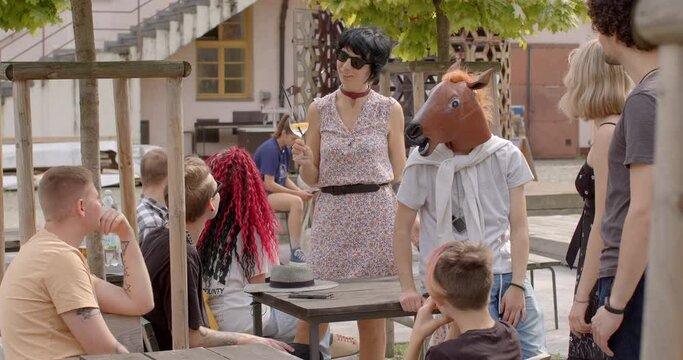 Young woman holding glass of non-alcoholic beverage joins group of young people. Her appearance is unconventional for teacher, reflecting sense of freedom in self-expression. Man wearing a horse mask