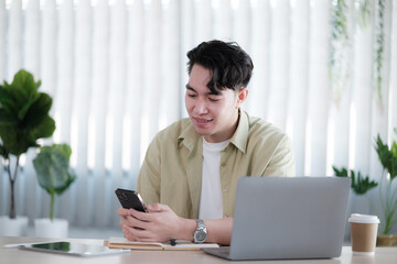 Young man reading sms message from friend on smartphone sitting with laptop device in co working...