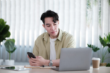 Young man reading sms message from friend on smartphone sitting with laptop device in co working space.