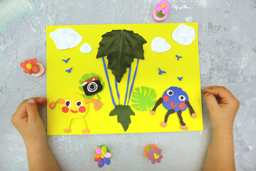 Child making card from paper, plasticine and natural leaves. Air balloon, clouds, birds, flowers......
