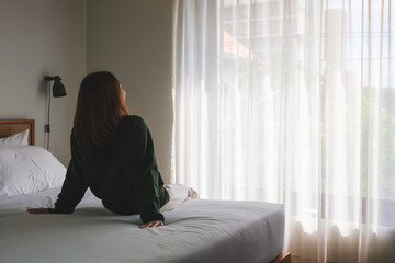 Rear view image of a woman sitting relaxing on a bed after waking up in bedroom - obrazy, fototapety, plakaty