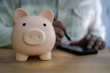 Pink piggy bank on wooden table and man writing and savings by smartphone, planning and future at...