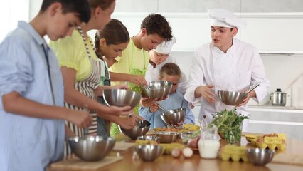 During lesson in cooking courses, male cook professional tells children about rules ticks and tricks for making fluffy pancakes, woman assistant helps hesitant child mix ingredients in bowl