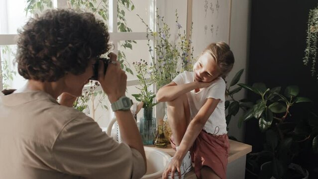 Modern preteen girl sitting on countertop in cozy kitchen posing on camera while her mom taking photos on camera