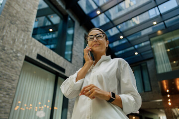 Smiling female sales manager talking phone with client while standing on coworking background