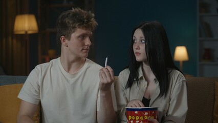 Portrait of teenage couple spending leisure time. Boy and girl sitting on the sofa, boy holds a cigarette joint and gives it to girl, girl looks confused.