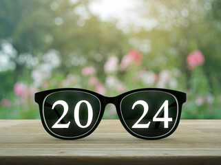 2024 white text with black eye glasses on wooden table over blur pink flower and tree in garden, Business vision happy new year 2024 cover concept
