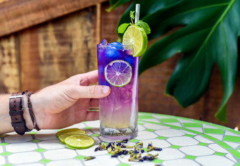 Hand holding lemonade with butterfly pea flowers and blue syrup on narutral leaves background with copy space