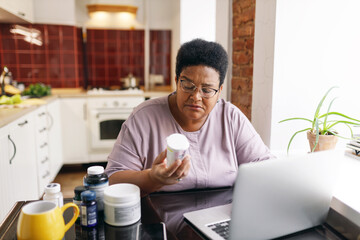 Serious plus size senior black female in glasses sitting at kitchen table full of food supplements using laptop, reading label on vitamin bottle, making online order on marketplace