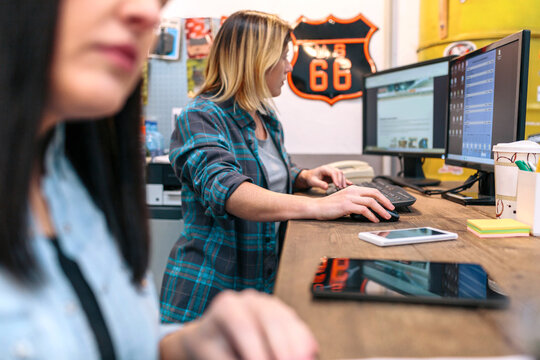 Blonde young woman checking orders in a computer received by ecommerce shop. Woman entrepreneur wearing a plaid shirt and working selling on line next to female colleague in small business.