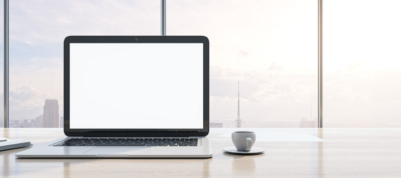 Close up of modern wooden desktop with clean white mock up laptop screen, supplies, coffee cup and panoramic window with city view in the background. 3D Rendering.