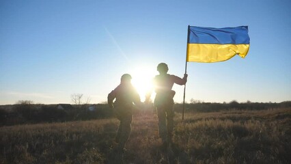 Military couple of ukrainian army holds hands of each other jogs with a waving Ukraine flag at meadow. Young soldiers in camouflage uniform running with lifted national banner on field at sunset