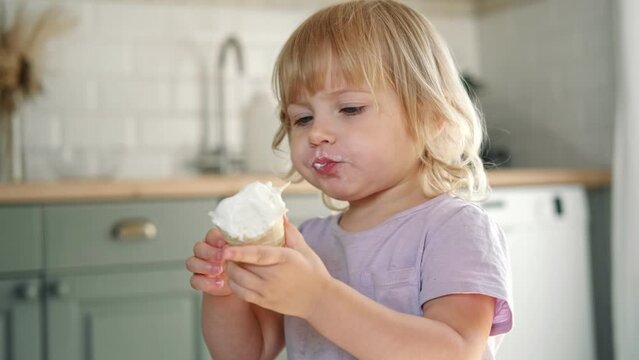 Baby girl enjoying ice cream. Pretty little toddler eating an ice-cream indoors, at home. Dining room background. Small child eats plombir and cream messy on her mouth. Cute kid with tasty sweet food.