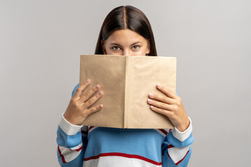 Brunette teen student with book in hands looking at camera on gray background. Hiding face behind...