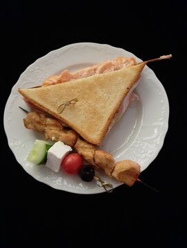 Triangular bread toast, skewers with pieces of fish and fresh vegetables with a cube of cheese all on a white plate. Party snacks on a white figured plate on a black background.