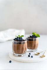 Delicious sweet chocolate mousse dessert in glasses decorated with chocolate cookie crumbs, fresh blueberry and mint on a light concrete background. Copy space. Confectionery menu, recipe.