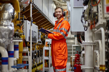 Portrait of young marine engineer in orange coverall doing daily check list in engine room. - obrazy, fototapety, plakaty