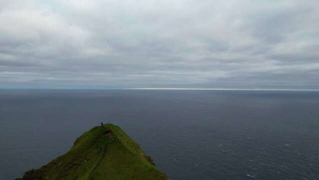 Aerial shot of adventurous hikers stand on a cliff edge taking pictures by Kallur lighthouse in Kalsoy, Faroe Islands