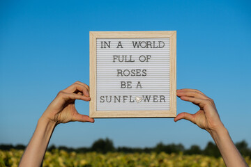 IN A WORLD FULL OF ROSES BE A SUNFLOWER text on white board next to sunflower field. Motivational...