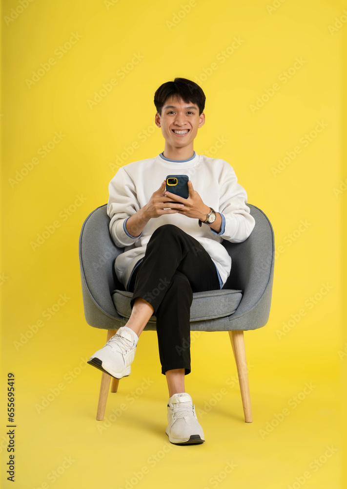 Wall mural full body of young Asian man wearing sweater and using phone on yellow background