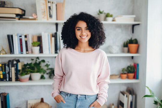 A beautiful biracial model woman smiling and standing in front of white shelves full of books and plants, black curly hair in light pink long sleeve sweatshirt with blue jean teen