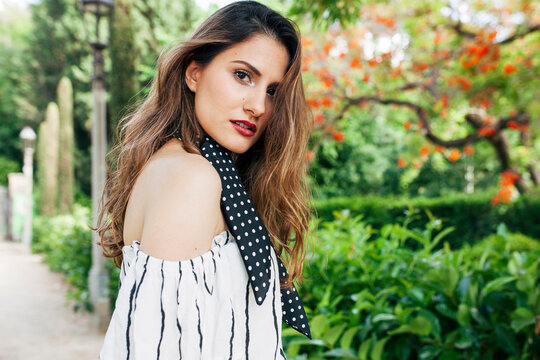 Relaxed young attractive elegant woman standing in park with a white dress and black scarf, looking at camera. 