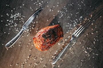 Juicy piece of ready meat lying on salt sprinkled on stone kitchen table next to kitchen utensils...