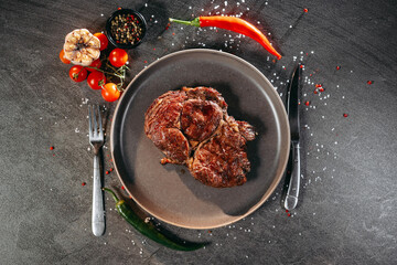 A large piece of grilled steak lying on a flat gray plate, next to which are cutlery fork and knife, as well as green and red hot peppers, a sprig of cherry tomatoes, a black jar of mixed peppercorns
