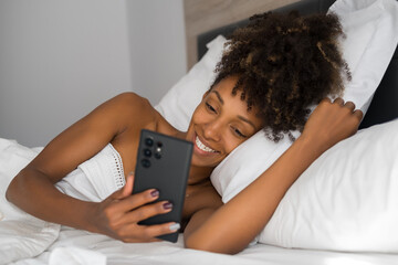 Portrait of a beautiful happy young black woman using smartphone in a comfortable bed in the morning. Afro hairstyle natural beauty holding her phone. Leisure, rest and relaxing at home.