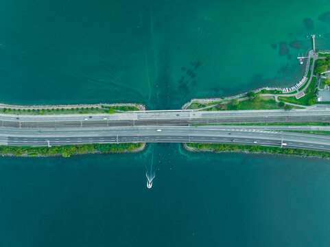 Aerial view of vehicles driving on a road crossing the Lugano lake with a bridge and a boat sailing under the bridge, Melide, Ticino, Switzerland.