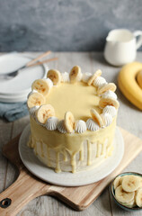 Banana and walnut cake with white chocolate ganache on the top, whipped cream and banana slices. Wooden chopping board and blue background.