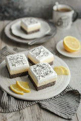 Creamy lemon and poppy seed bars on a white plate decorated with fresh lemon slices. Grey background and stripped napkin. Cup of coffee.