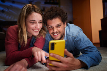Happy nice Caucasian couple using cell phone smiling lying on carpet in living room at home at...