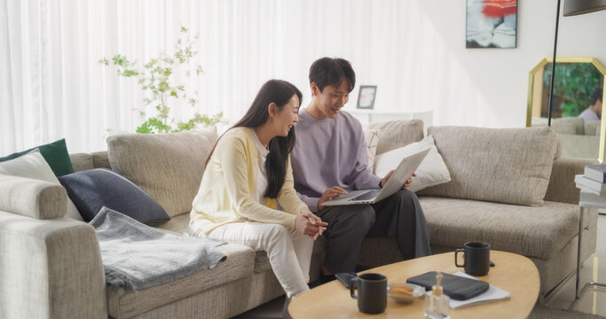 Diverse South Korean Couple Cherishing Moments Together, Using Laptop Computer on a Couch of Their Bright and Comfortable Living Room. Young Man and Woman Designing Online Invitation to a Party