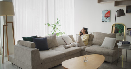 Sitting on a Couch in Her Cozy Modern Living Room, Beautiful Smiling Korean Woman is Having a Phone Conversation, Talking with Girlfriend about Latest Fashion Trends and Favorite TV Show Characters