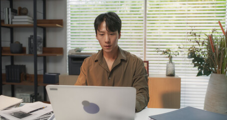 Young and Handsome South Korean Professional, Immersed in Laptop Computer Work Behind a Desk at His...