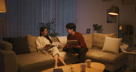 Young and Loving Asian Couple Relaxing at Home. South Korean Partners Sitting on a Couch, Female Reading a Trendy Magazine, Male Using a Tablet Computer and Spending Time Online on Social Media App