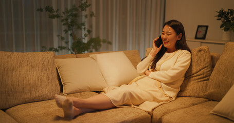 Obraz premium Relaxing on a Couch in the Evening in a Cozy Apartment, Beautiful Smiling South Korean Woman is Having a Funny Phone Conversation with Friend, Sharing Happy Stories About Relationships and Friendship 