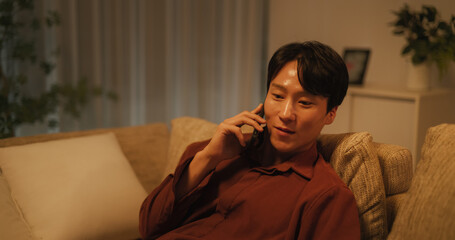 Fototapeta premium Excited South Korean Man Answering a Phone Call. Handsome Asian Male Sits Comfortably on the Sofa in His Cozy Living Room in The Evening, Talking with Friends, Family or Loved One 